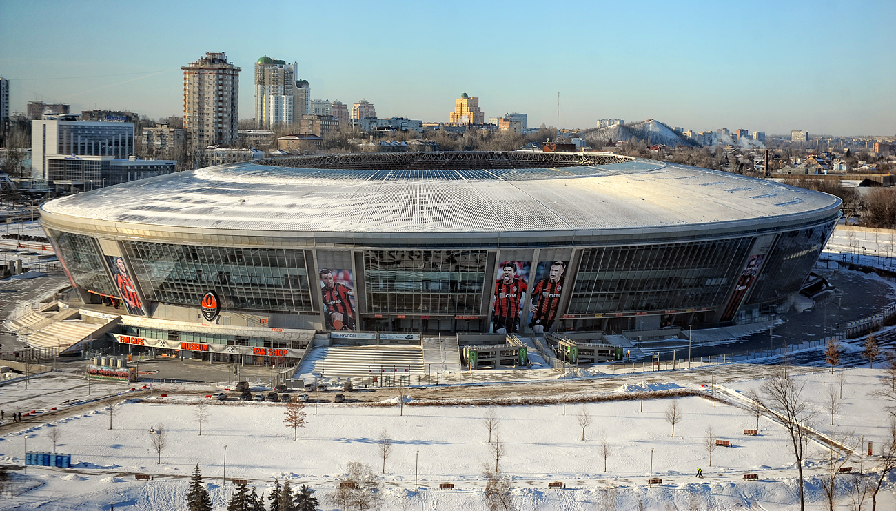 Winter aerial view of the Donbass Arena in Donetsk showing the oval stadium with a snow-covered roof and the surrounding cityscape.