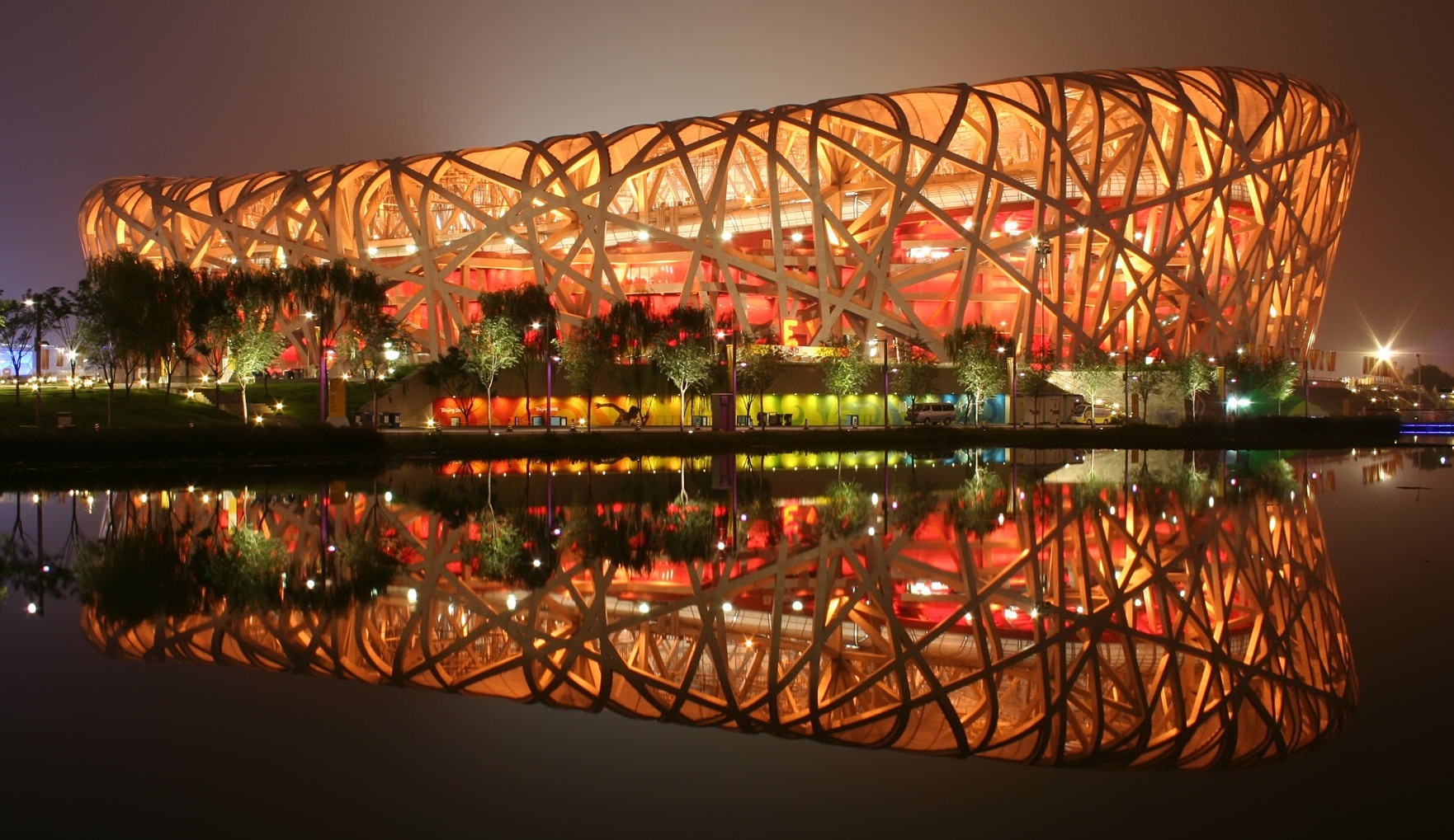 Night view of the Birds Nest Stadium in Beijing, illuminated in warm orange light with its steel structure reflected in still water in the foreground