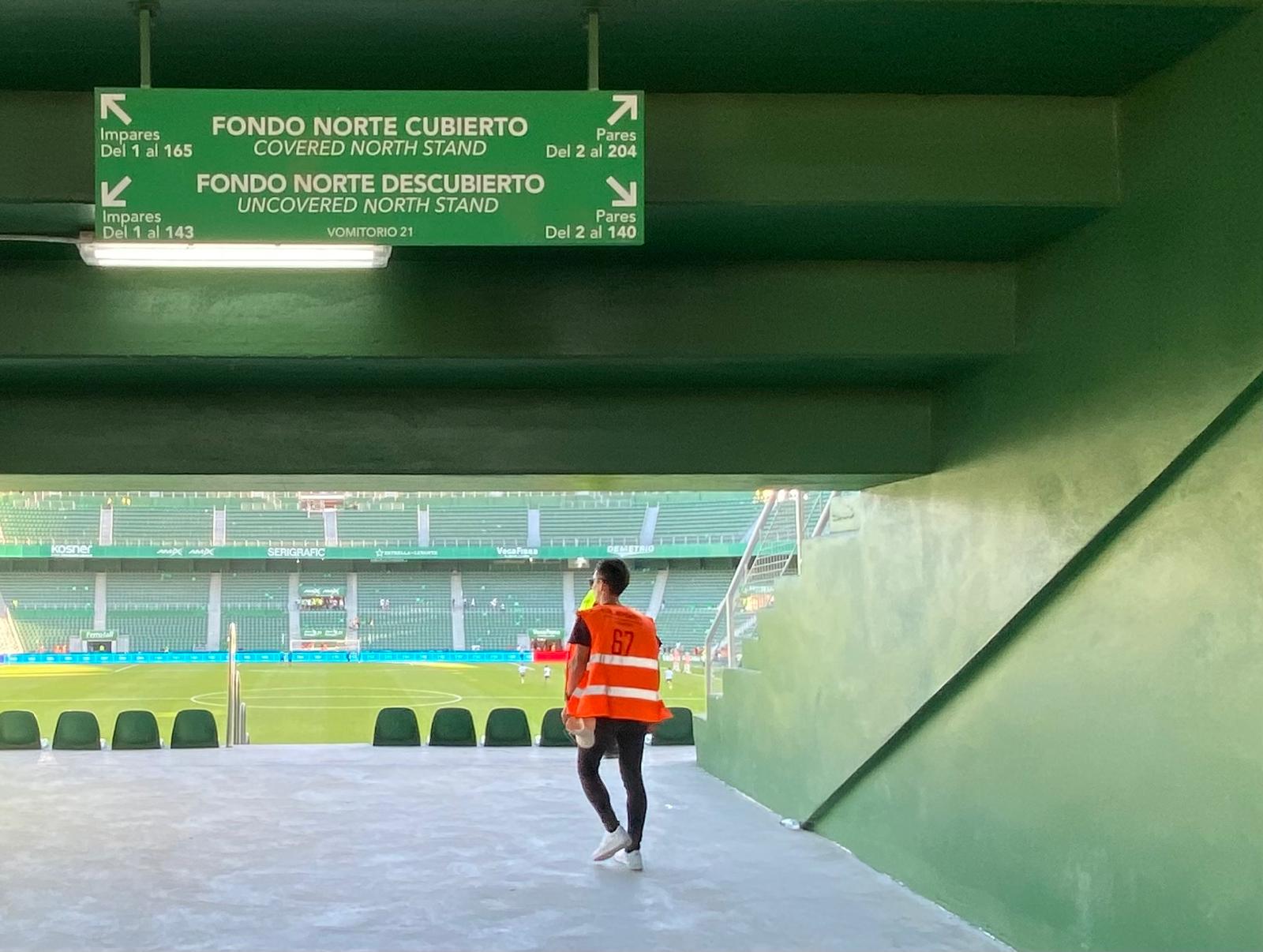 A stadium steward in an orange vest walks through a green concrete tunnel toward the pitch at Estadio Martínez Valero, with signage for the covered and uncovered north stand visible above and empty green seats in the background.