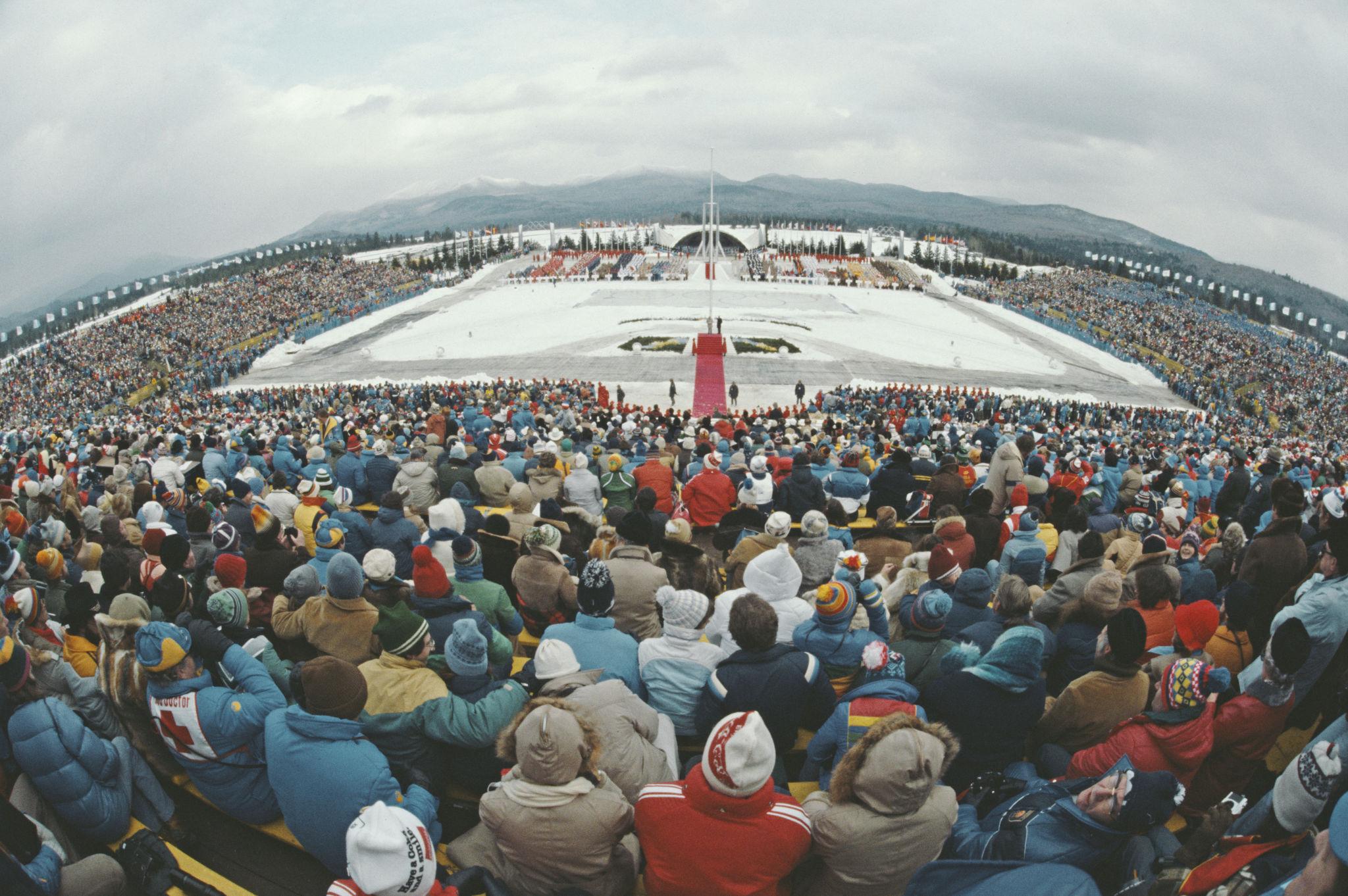 Wide view of the opening ceremony at the 1980 Winter Olympics in Lake Placid, with packed stands surrounding a snow covered stadium set against the Adirondack Mountains, highlighting the scale of the event within a small mountain setting.