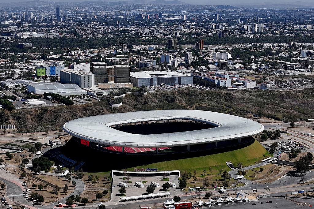 Aerial view of Estadio Akron Guadalajara showing the grass covered stadium structure and the city skyline of Guadalajara in the background.