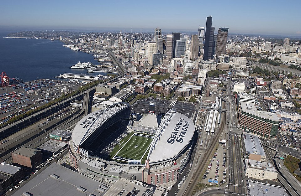 Aerial view of Lumen Field in Seattle, with the stadium beside downtown, rail lines, parking lots and the waterfront.
